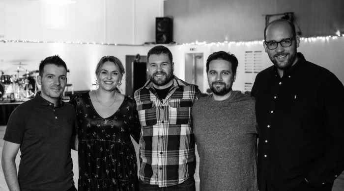 Black and white photo of five people standing close together indoors, smiling at the camera. They are in a casual setting with string lights in the background and music equipment visible on the left.